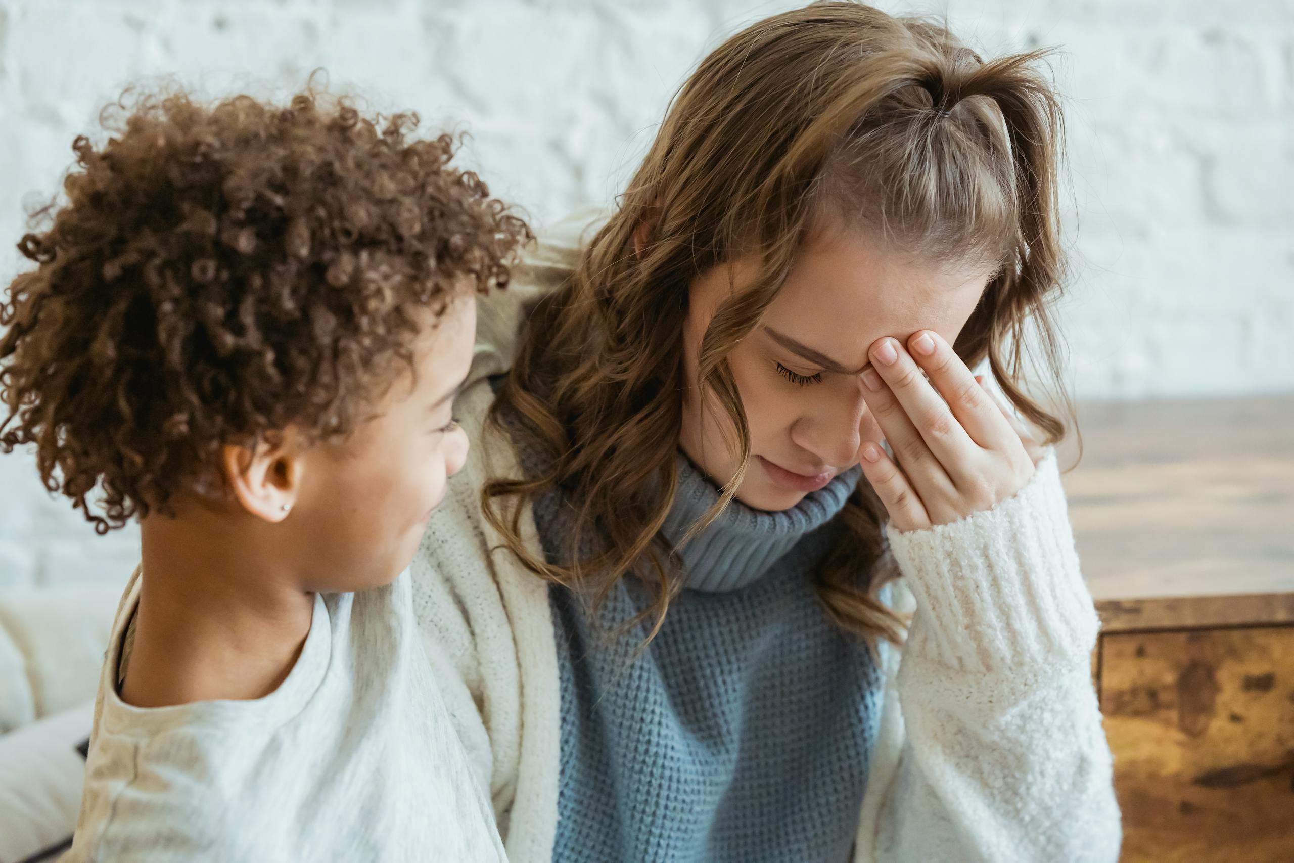 Cheerful African American son with curly hair consoling unhappy mother with closed eyes sitting in light room at home near white wall