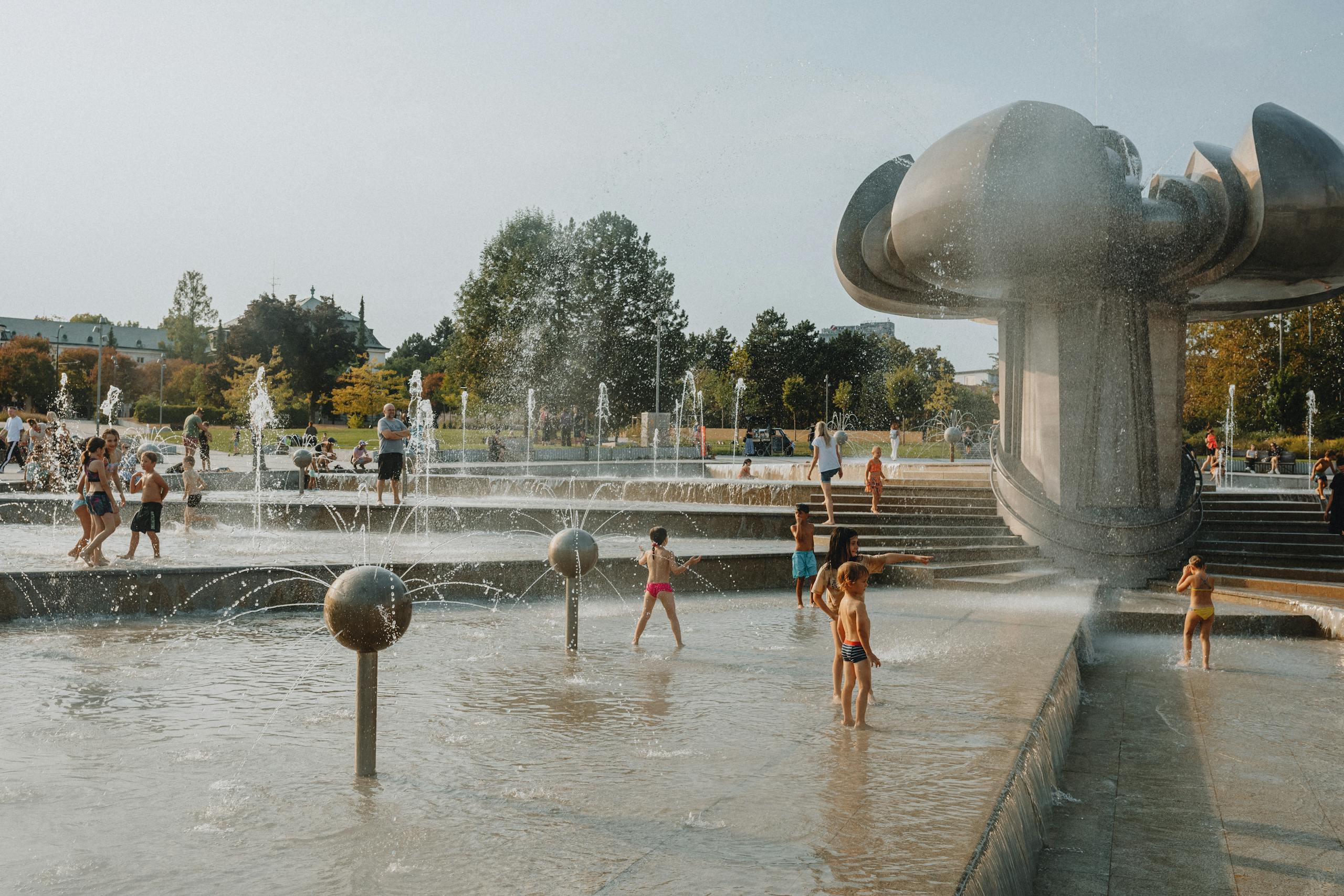 Children and adults enjoy a sunny day at a city park fountain, creating a vibrant, playful scene.