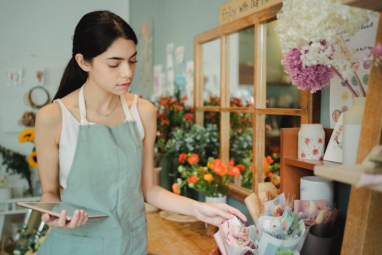 Concentrated female florist with dark hair standing in floral shop with tablet near flowers in wrapper while checking order during work