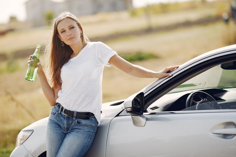 Confident female driver leaning on car and looking at camera while drinking water from glass bottle against blurred background in sunny summer day