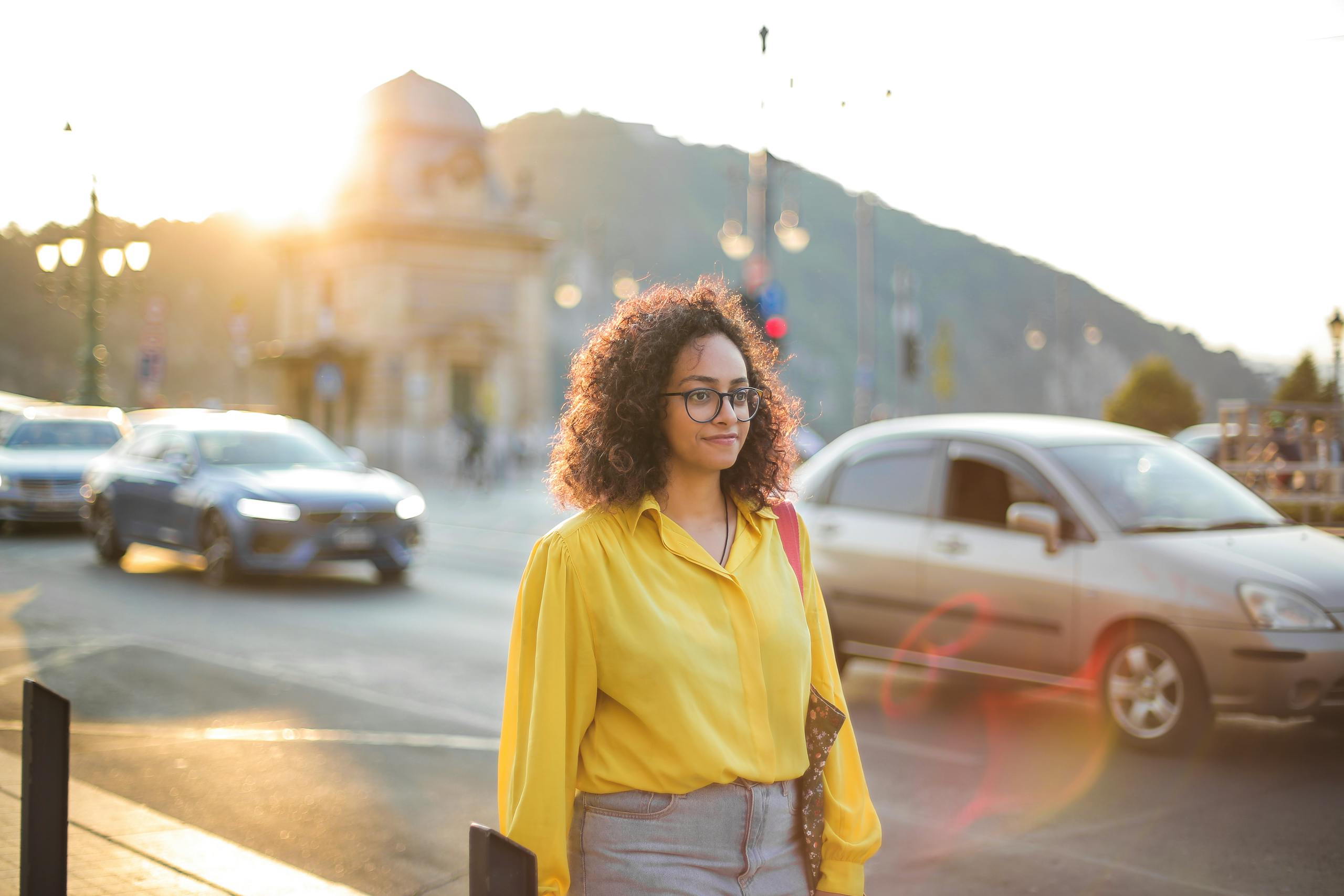 Confident woman in yellow shirt walks along a busy city street at sunset, with cars passing by.