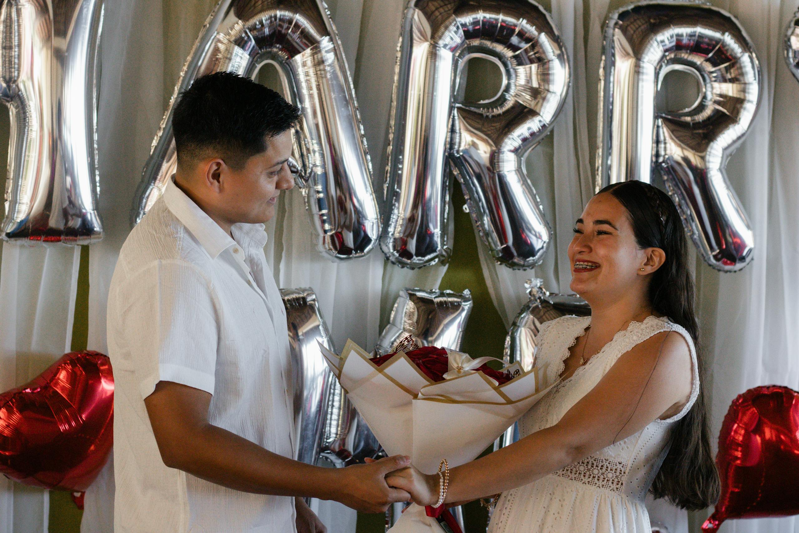 Couple celebrating engagement with balloons and bouquet indoors. Joyful and festive atmosphere captured.