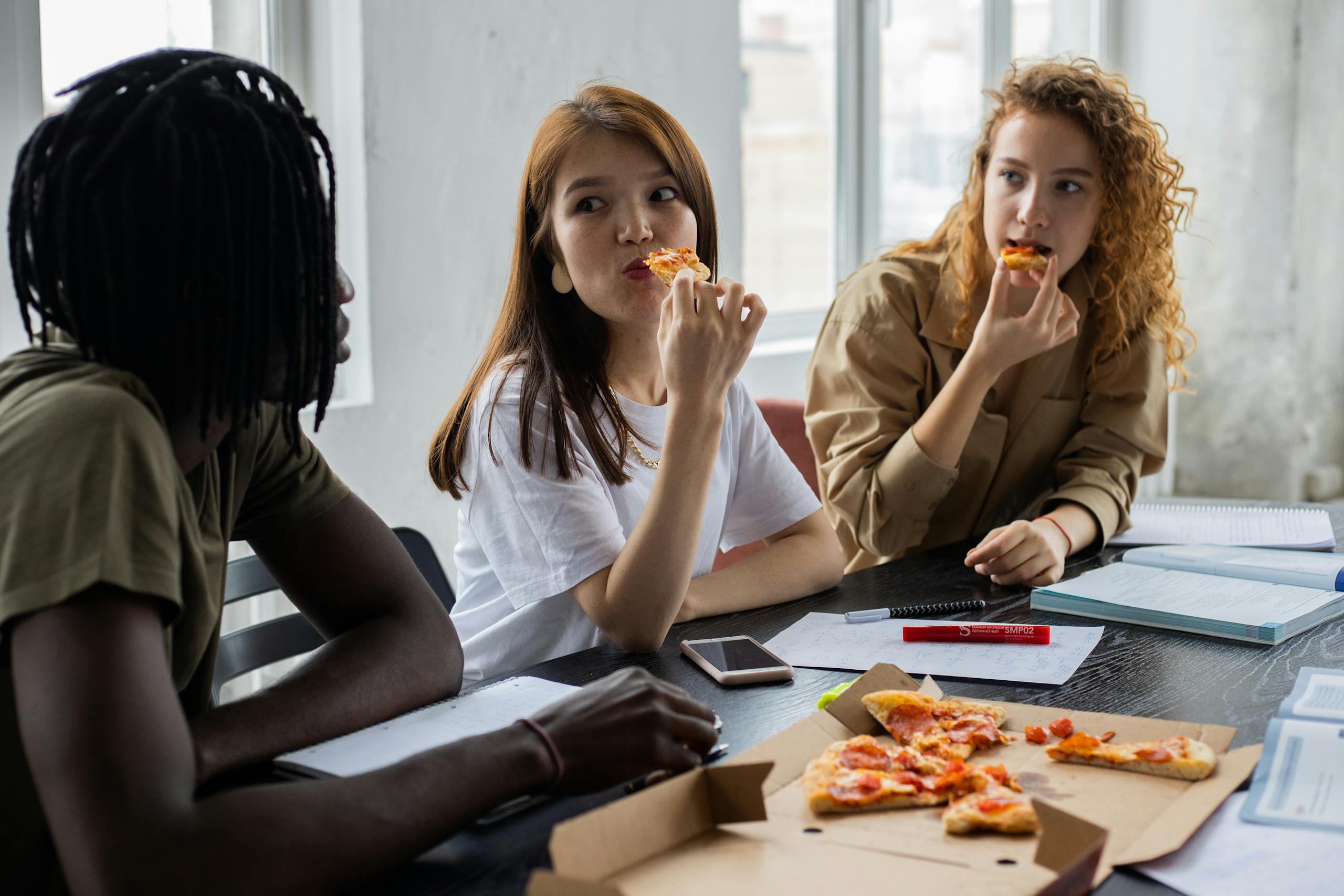 Diverse classmates sitting at table with gadget and textbooks and eating delivered pizza and at lunch together