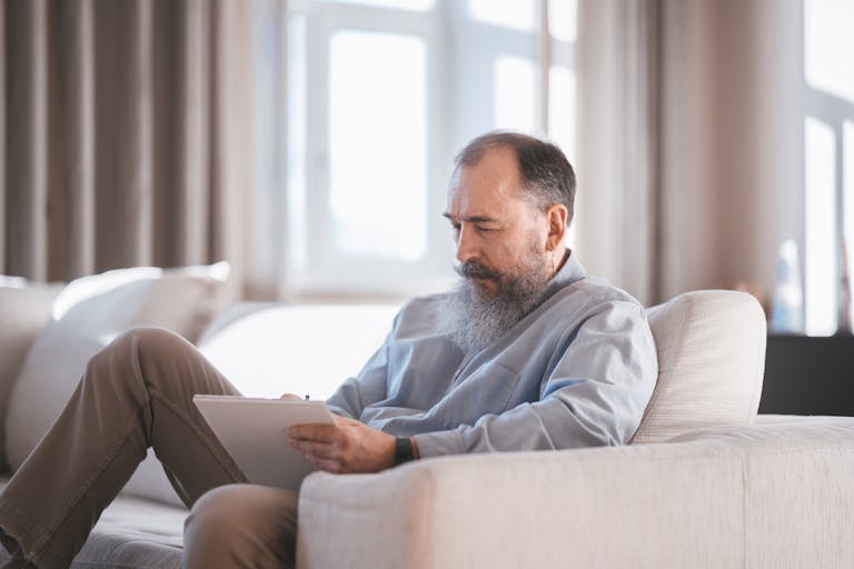 Elderly man sitting on sofa writing on a tablet with focus and concentration.