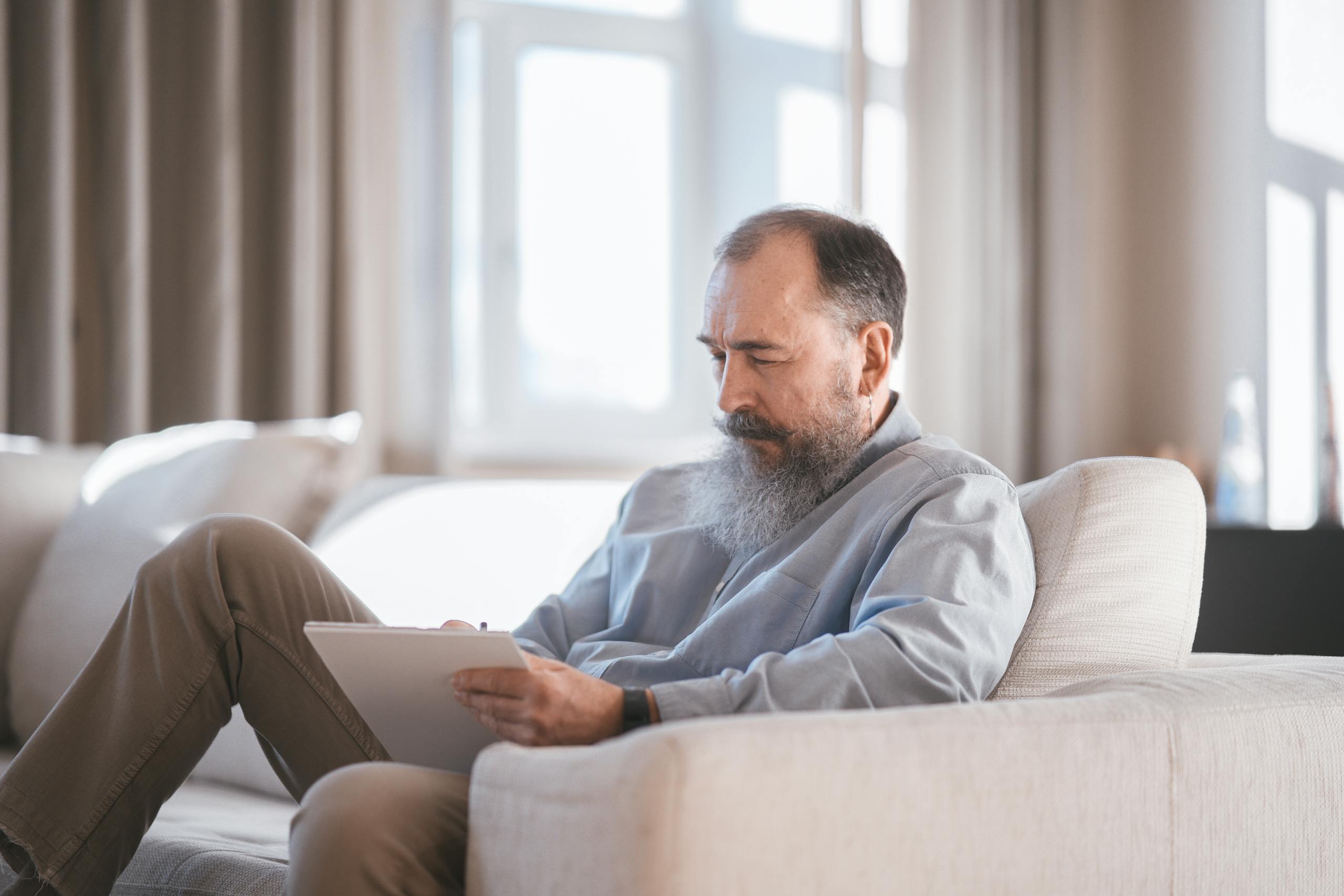 Elderly man sitting on sofa writing on a tablet with focus and concentration.