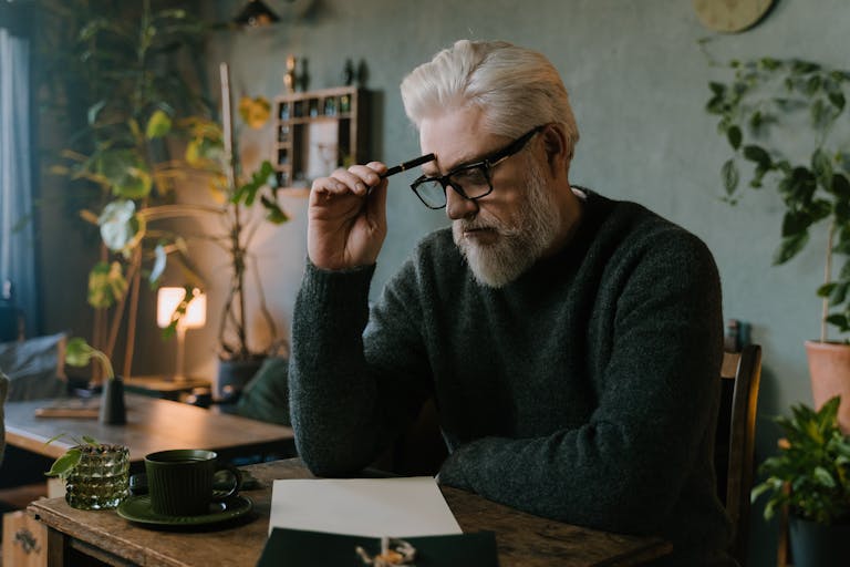 Elderly man with gray hair and beard thinking while sitting at table.