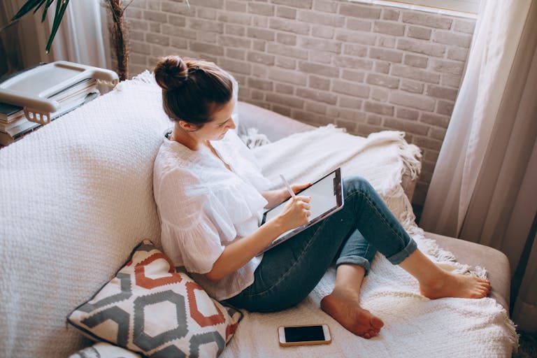 From above of young focused female in casual clothes sitting on sofa and drawing illustrations in tablet at home