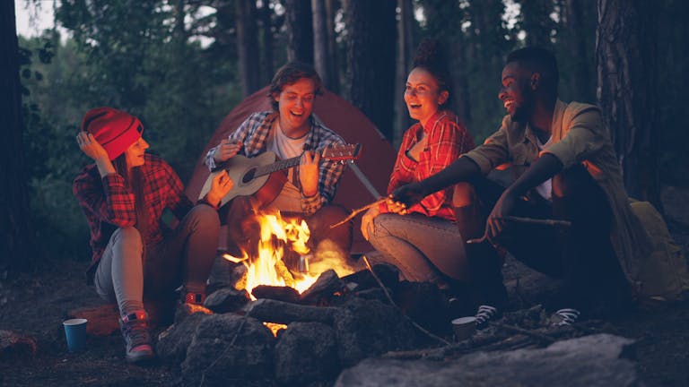 Group of friends camping, playing guitar and enjoying campfire in the woods at night.