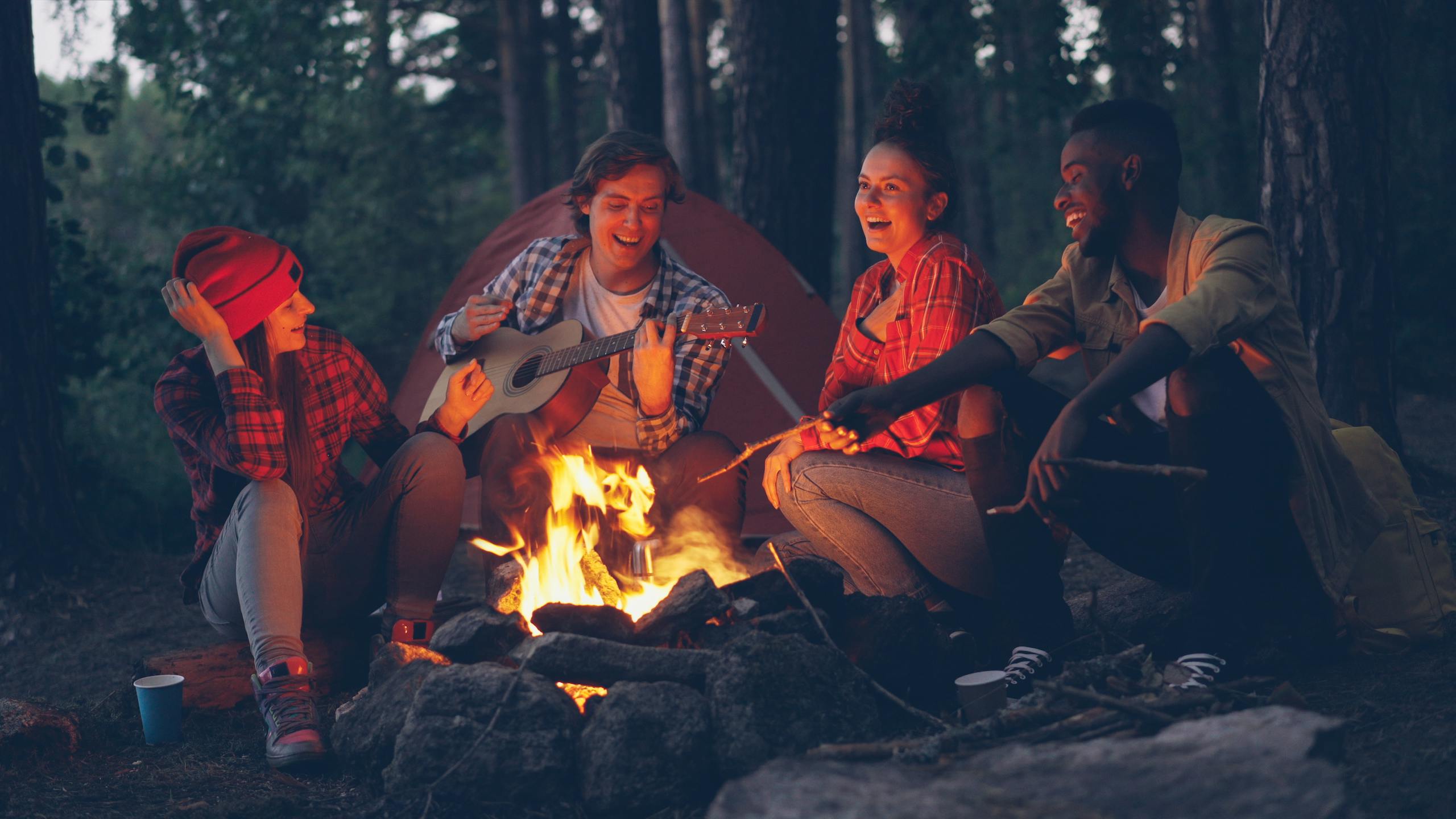 Group of friends camping, playing guitar and enjoying campfire in the woods at night.