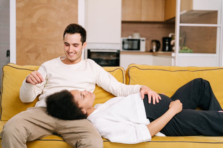 Happy interracial couple relaxing on a yellow sofa, embracing pregnancy moments together.