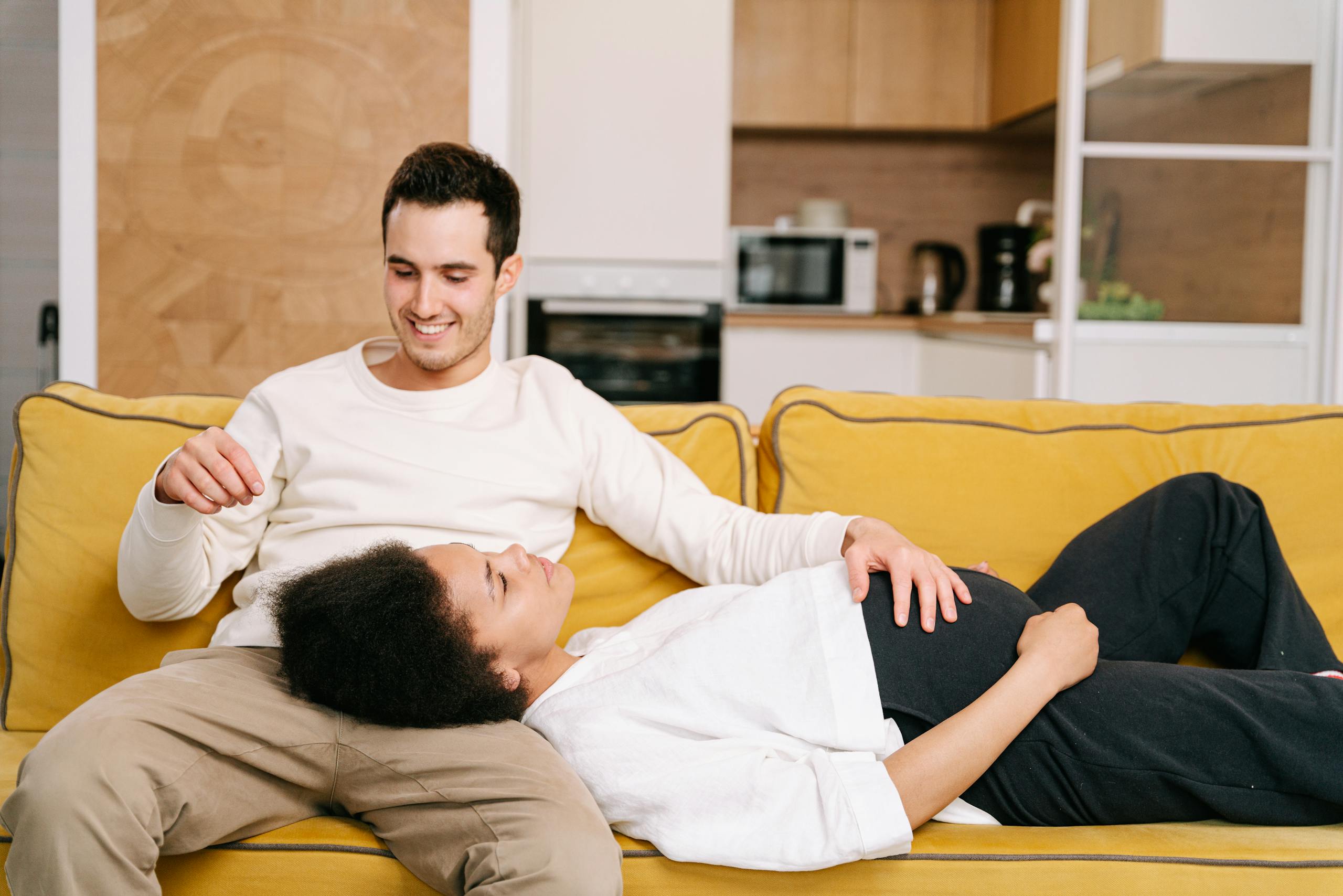 Happy interracial couple relaxing on a yellow sofa, embracing pregnancy moments together.