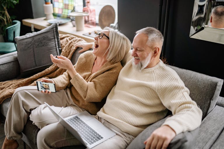 Happy senior couple laughing together while sitting on a cozy sofa with a laptop and smartphone.