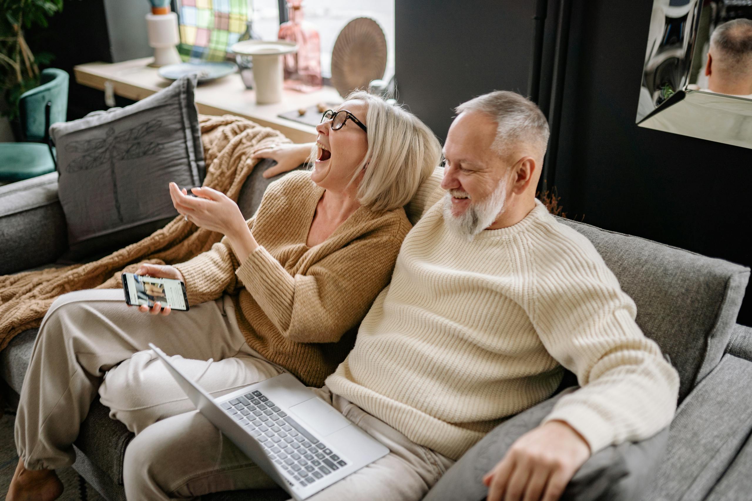 Happy senior couple laughing together while sitting on a cozy sofa with a laptop and smartphone.