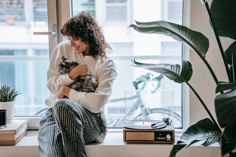 Happy young female in striped trousers embracing spotty cute fluffy cat on window in daytime