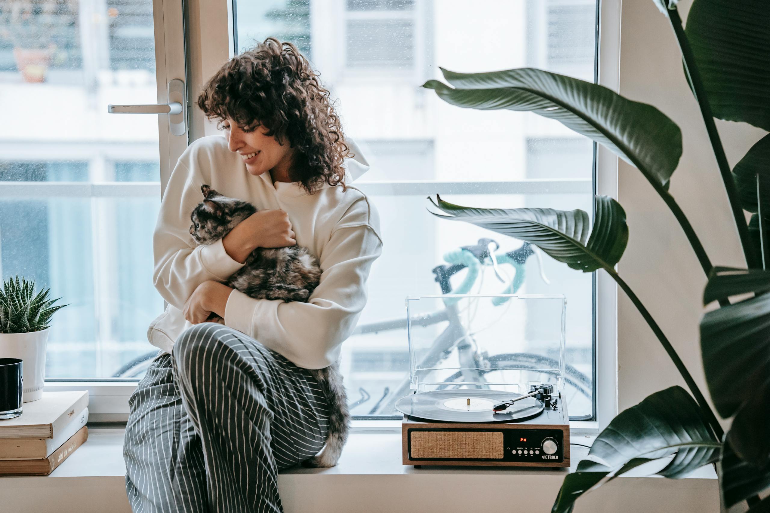 Happy young female in striped trousers embracing spotty cute fluffy cat on window in daytime