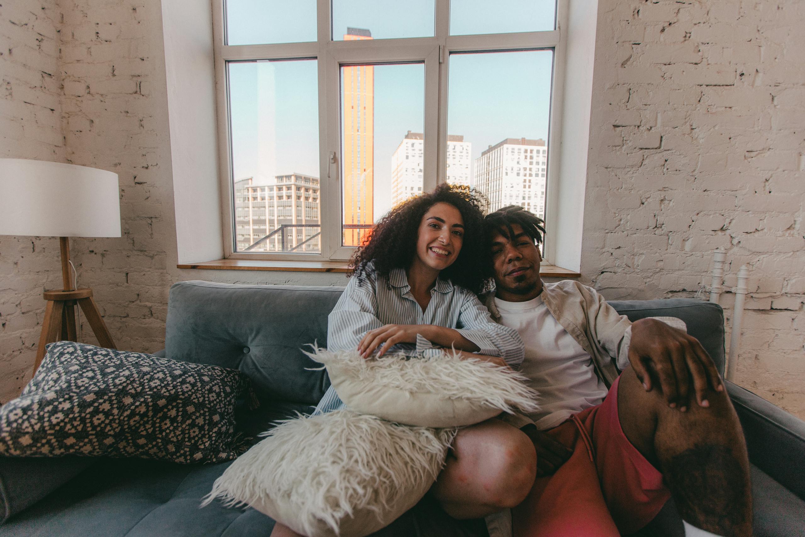 Joyful couple sitting together on a sofa in a modern apartment, with city views framing their warm smiles.