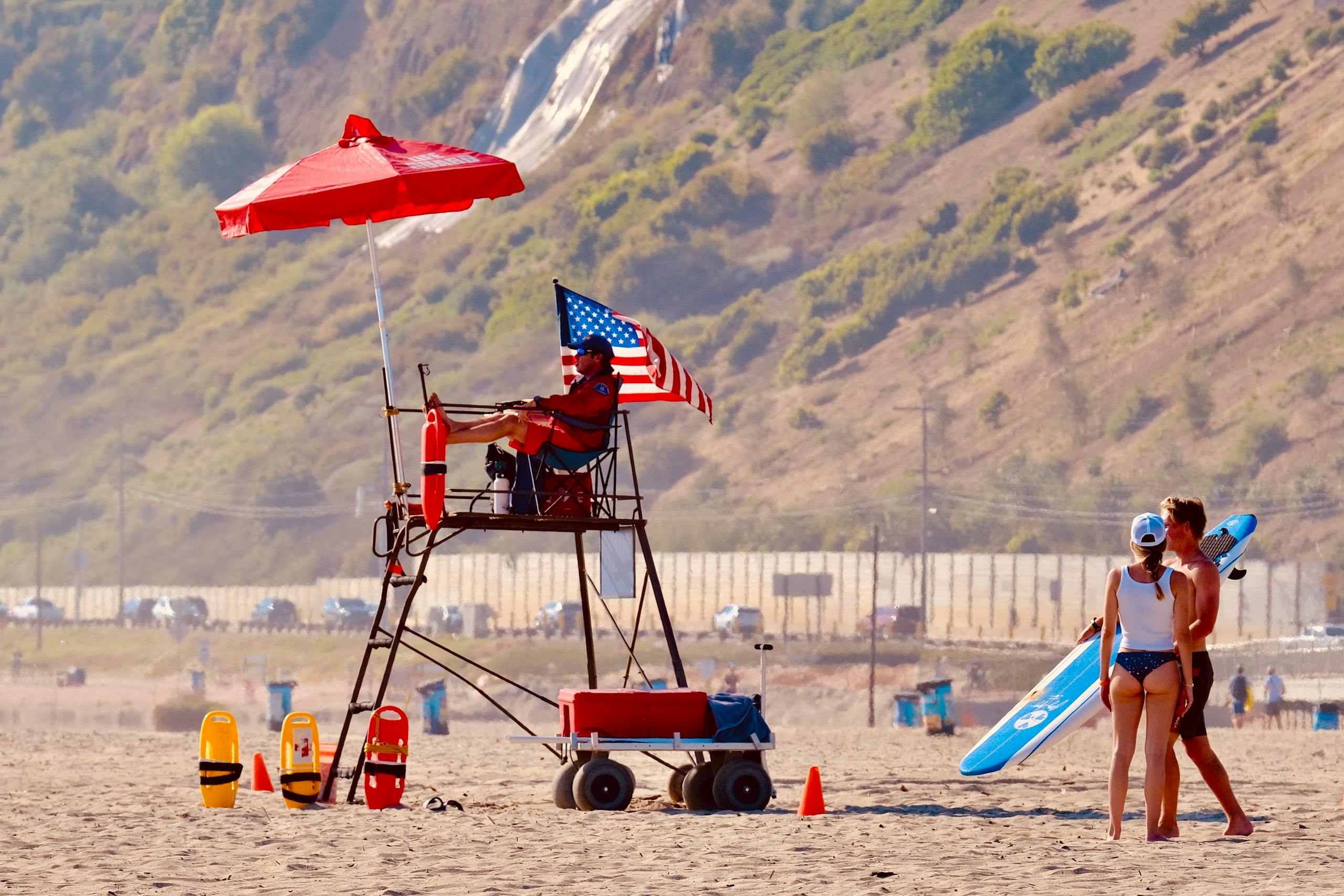 Lifeguard tower on a sandy beach with an American flag, overlooking people enjoying the sun.