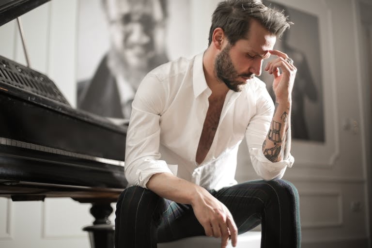 Low angle of handsome bearded man in white shirt touching forehead in deep thoughts sitting at piano