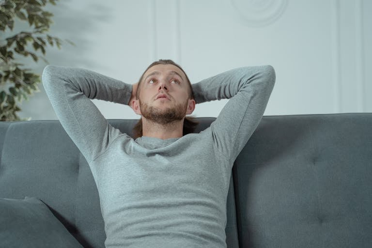 Man looking thoughtful while relaxing on a grey sofa indoors, conveying a sense of contemplation and relaxation.