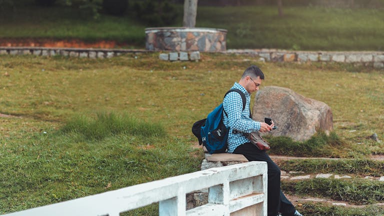 Man sitting in a park with a backpack and cellphone, enjoying a quiet moment.