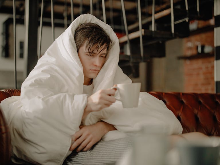 Man wrapped in a blanket sipping hot drink while seated on a cozy sofa indoors.