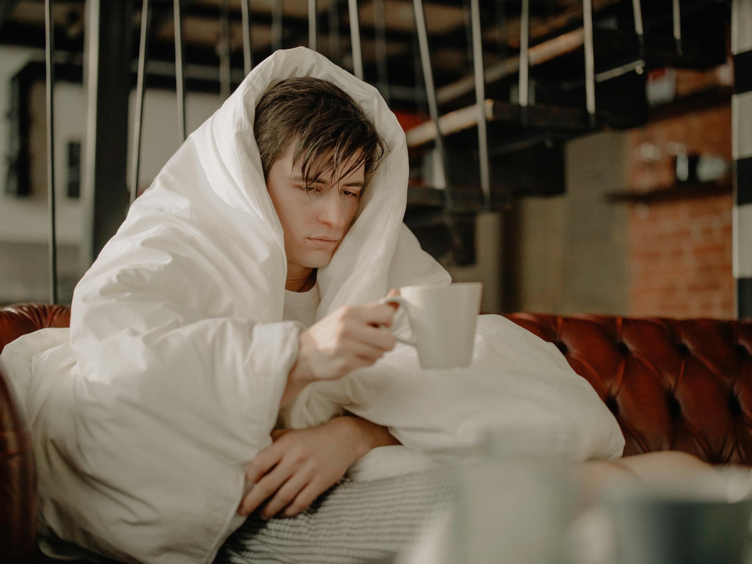 Man wrapped in a blanket sipping hot drink while seated on a cozy sofa indoors.