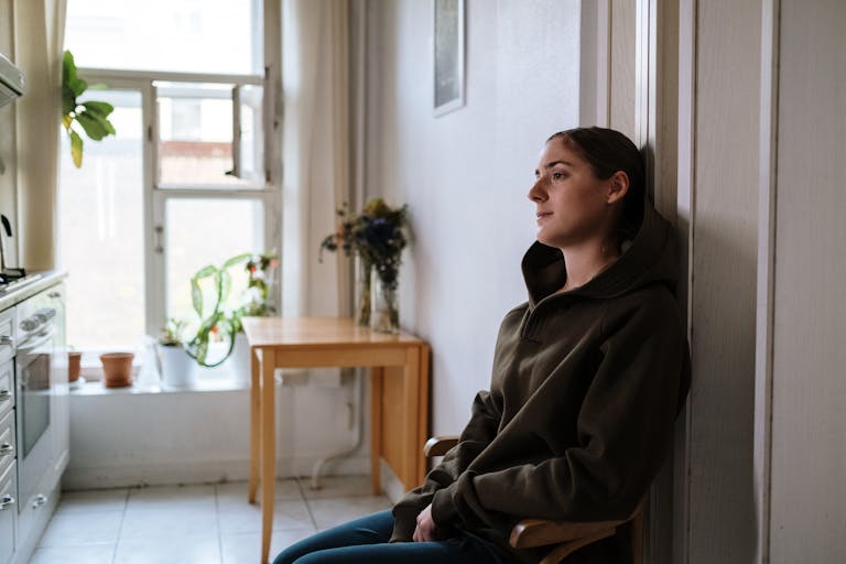 Pensive woman in a hoodie sitting indoors by a window, capturing a moment of reflection.