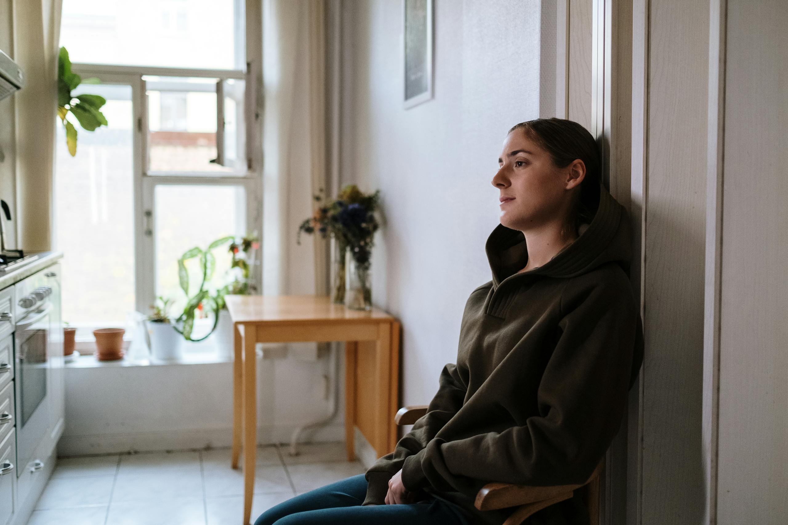 Pensive woman in a hoodie sitting indoors by a window, capturing a moment of reflection.
