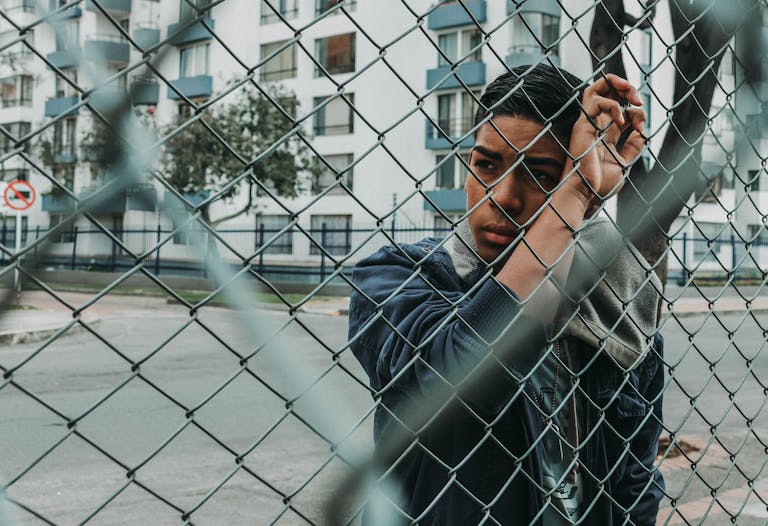 Pensive young man stands behind a chain-link fence in an urban setting, evoking a sense of contemplation.