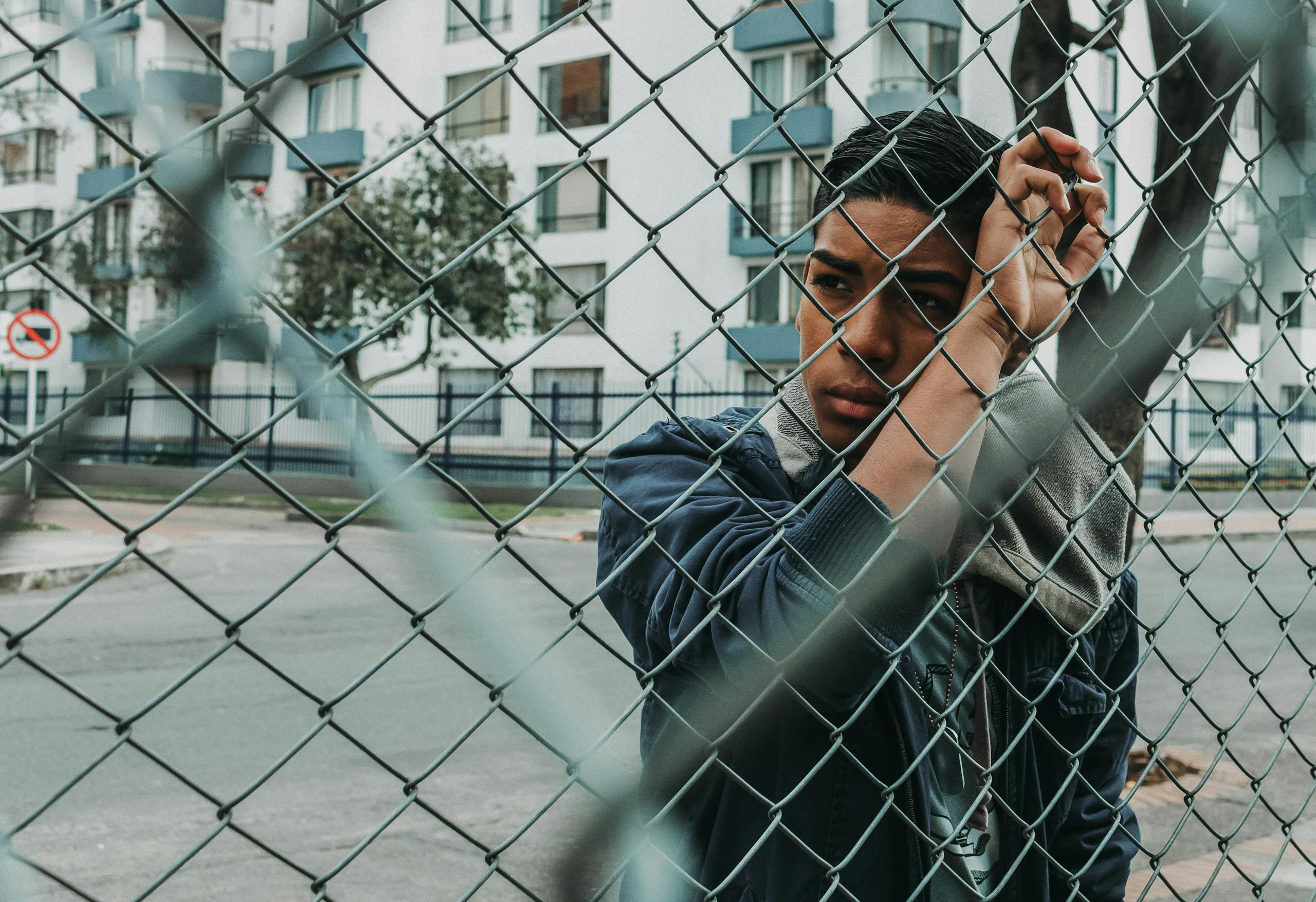 Pensive young man stands behind a chain-link fence in an urban setting, evoking a sense of contemplation.