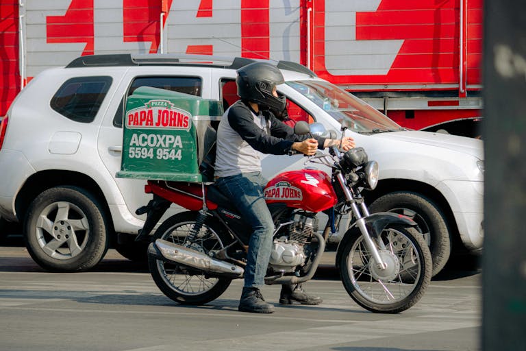 Pizza delivery rider on a motorcycle waits at a busy city street intersection.