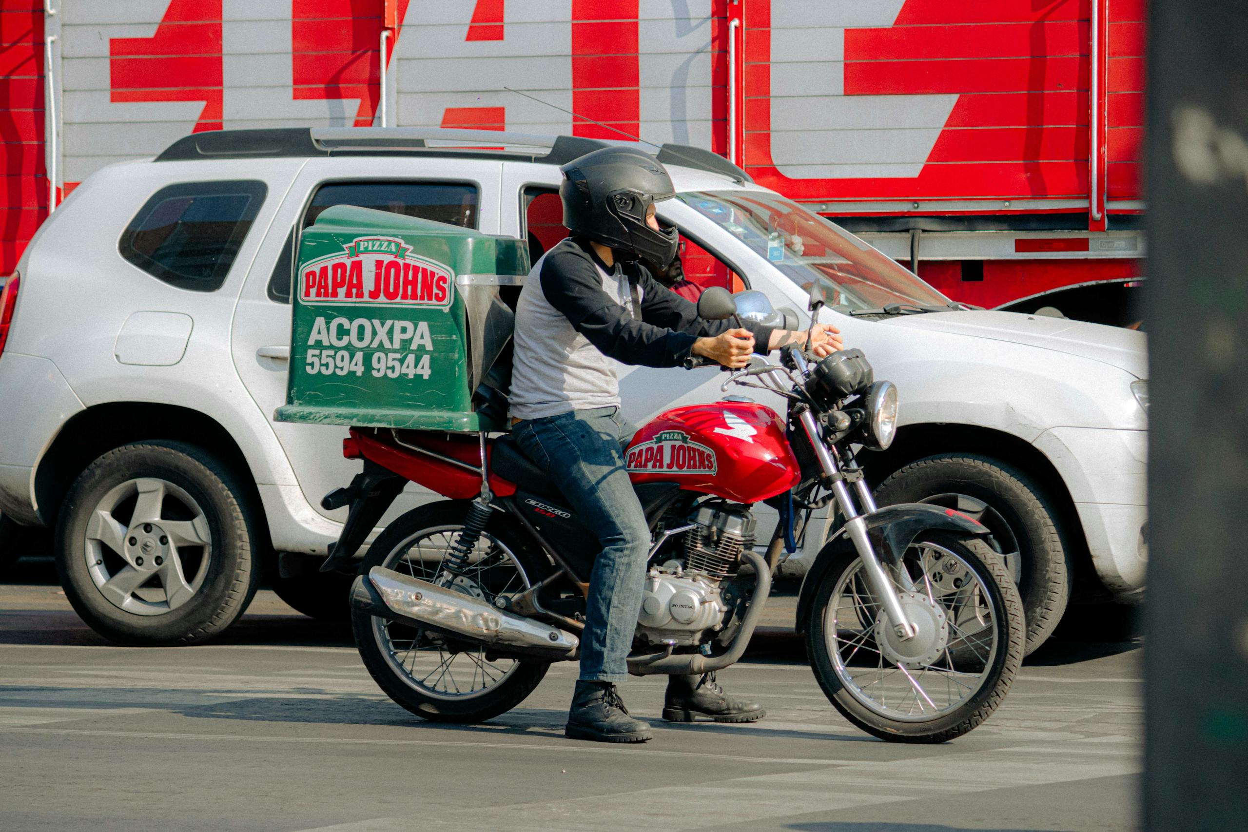 Pizza delivery rider on a motorcycle waits at a busy city street intersection.