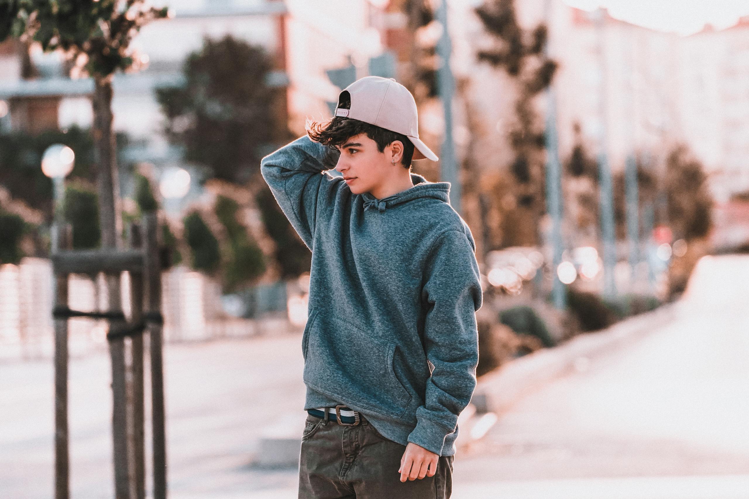 Pondering male teen in stylish wear touching cap on pavement in town while looking away