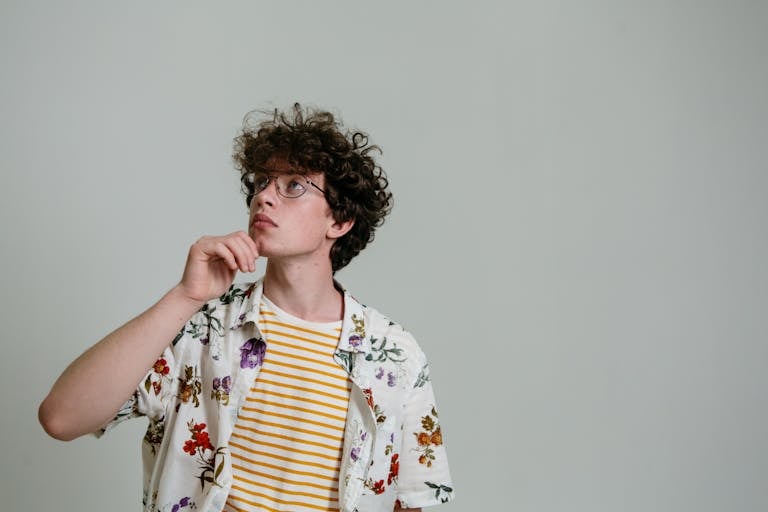 Portrait of a stylish young man posing thoughtfully in a studio setting with glasses and floral shirt.