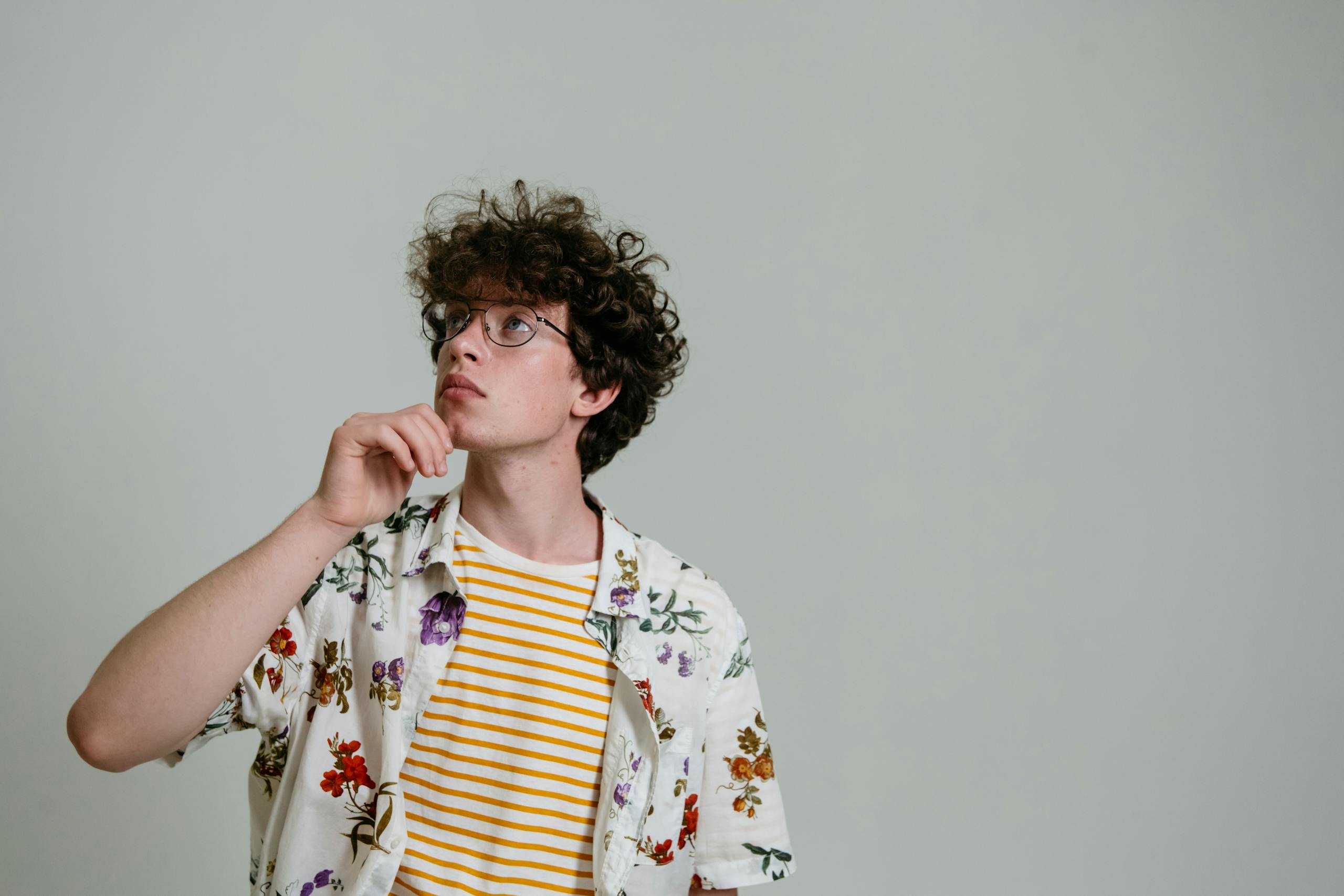 Portrait of a stylish young man posing thoughtfully in a studio setting with glasses and floral shirt.