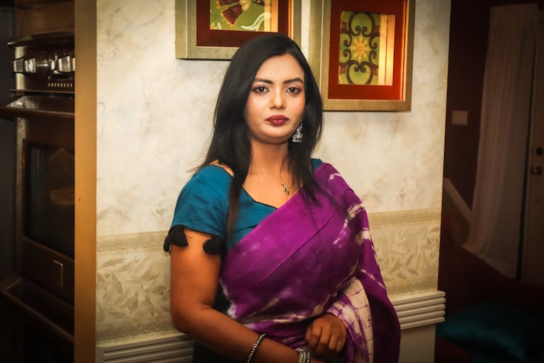 Portrait of an elegant woman in a traditional sari, standing indoors by decorated walls.