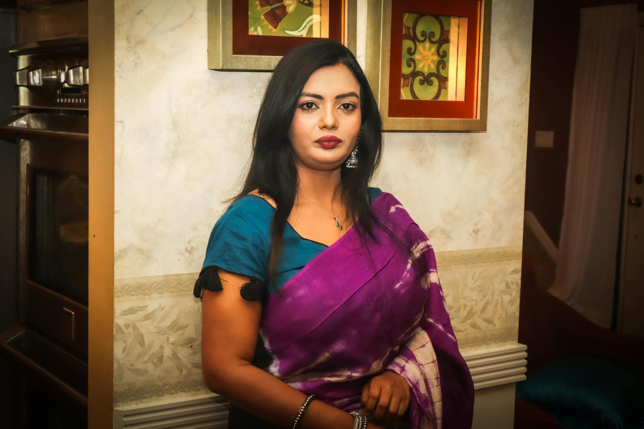 Portrait of an elegant woman in a traditional sari, standing indoors by decorated walls.