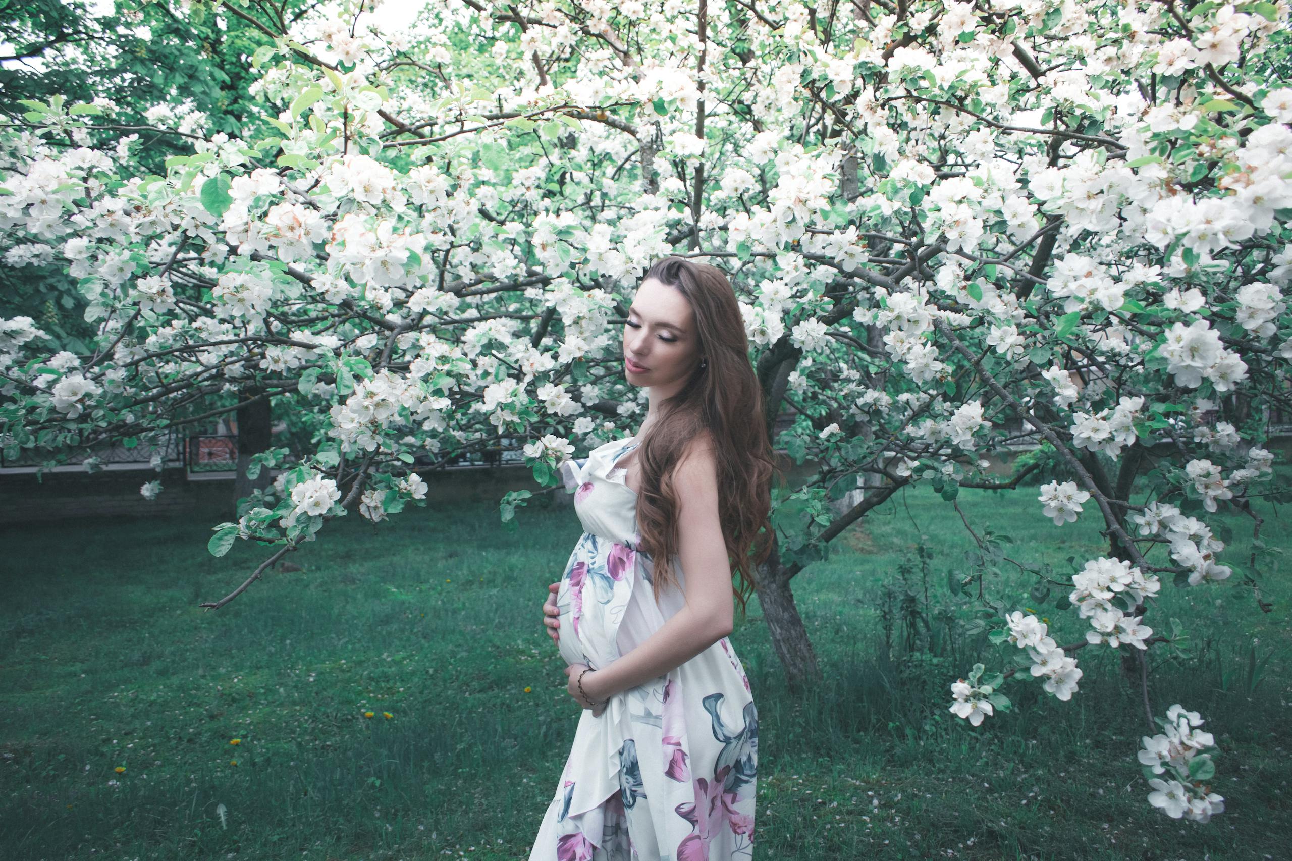 Pregnant woman in a floral dress, embracing spring blooms in an orchard. Captures serenity and new beginnings.