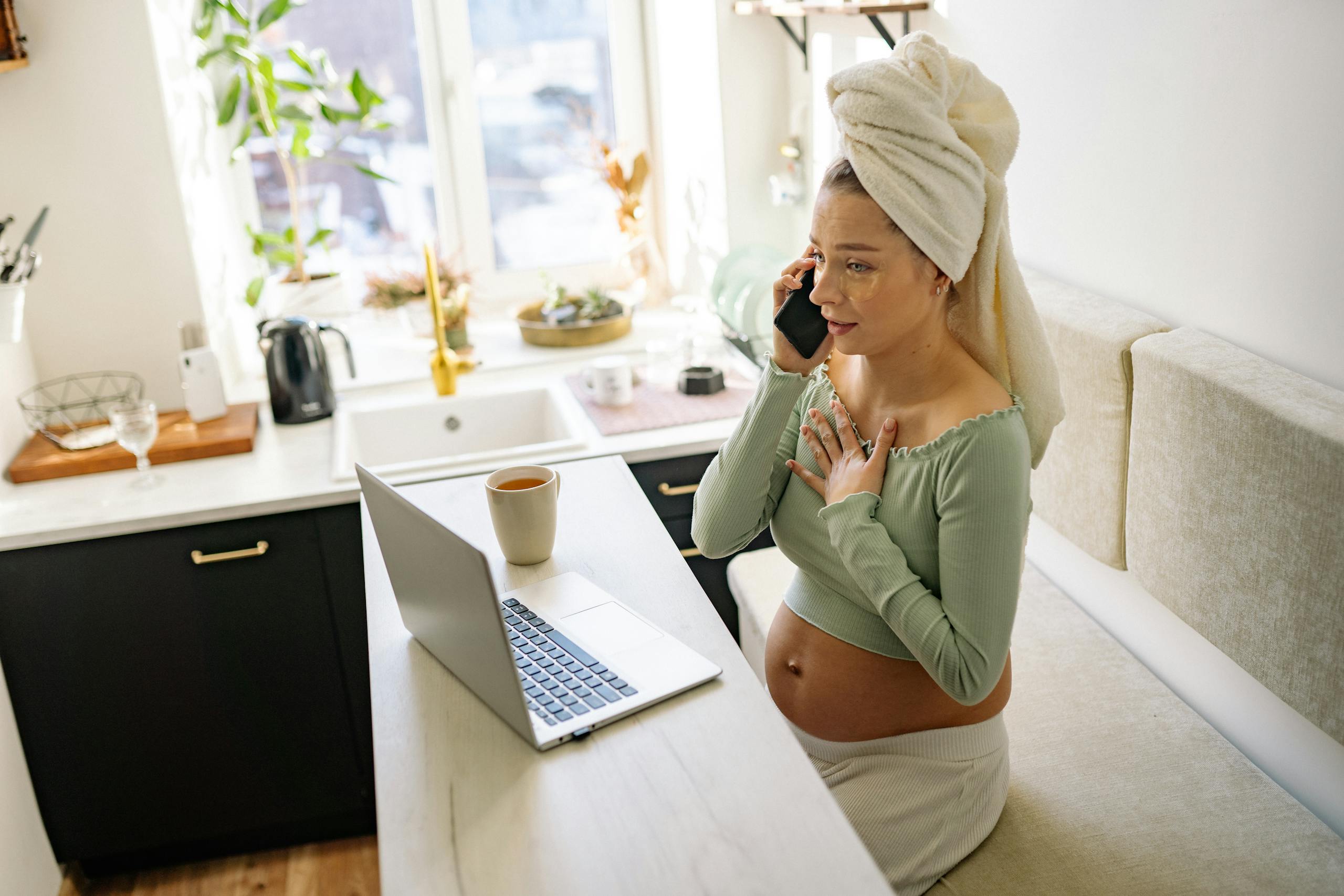 Pregnant woman in casual setting, talking on phone with laptop open and a drink nearby.