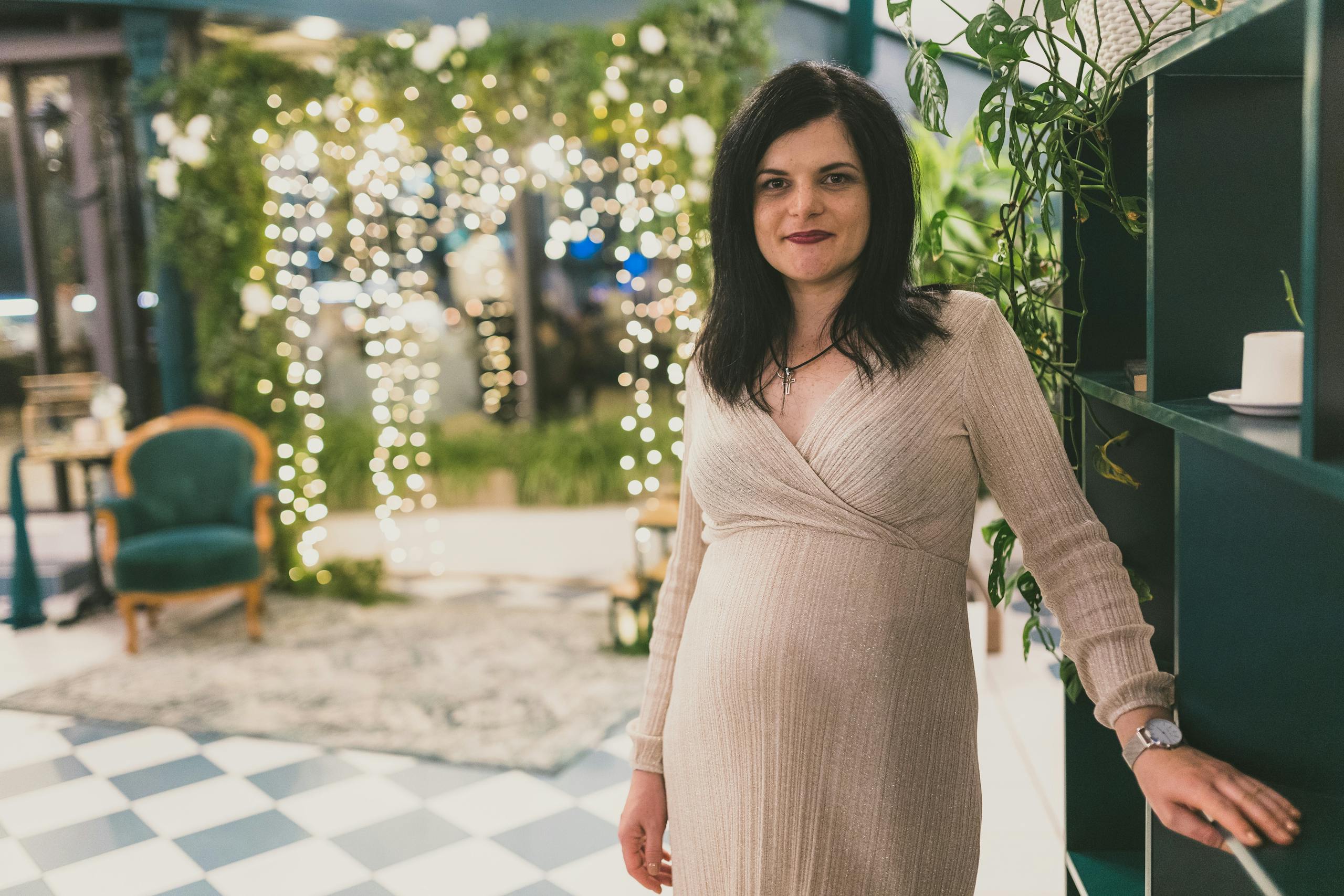 Pregnant woman in elegant dress standing indoors with soft lighting and greenery.