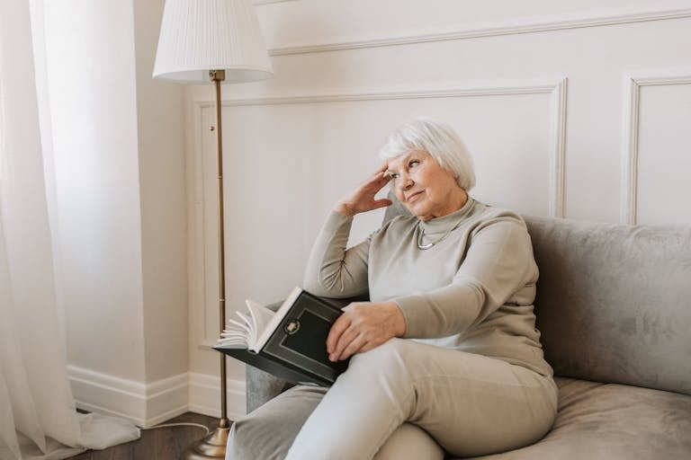Senior woman sitting on a couch reading a book, with a thoughtful expression.