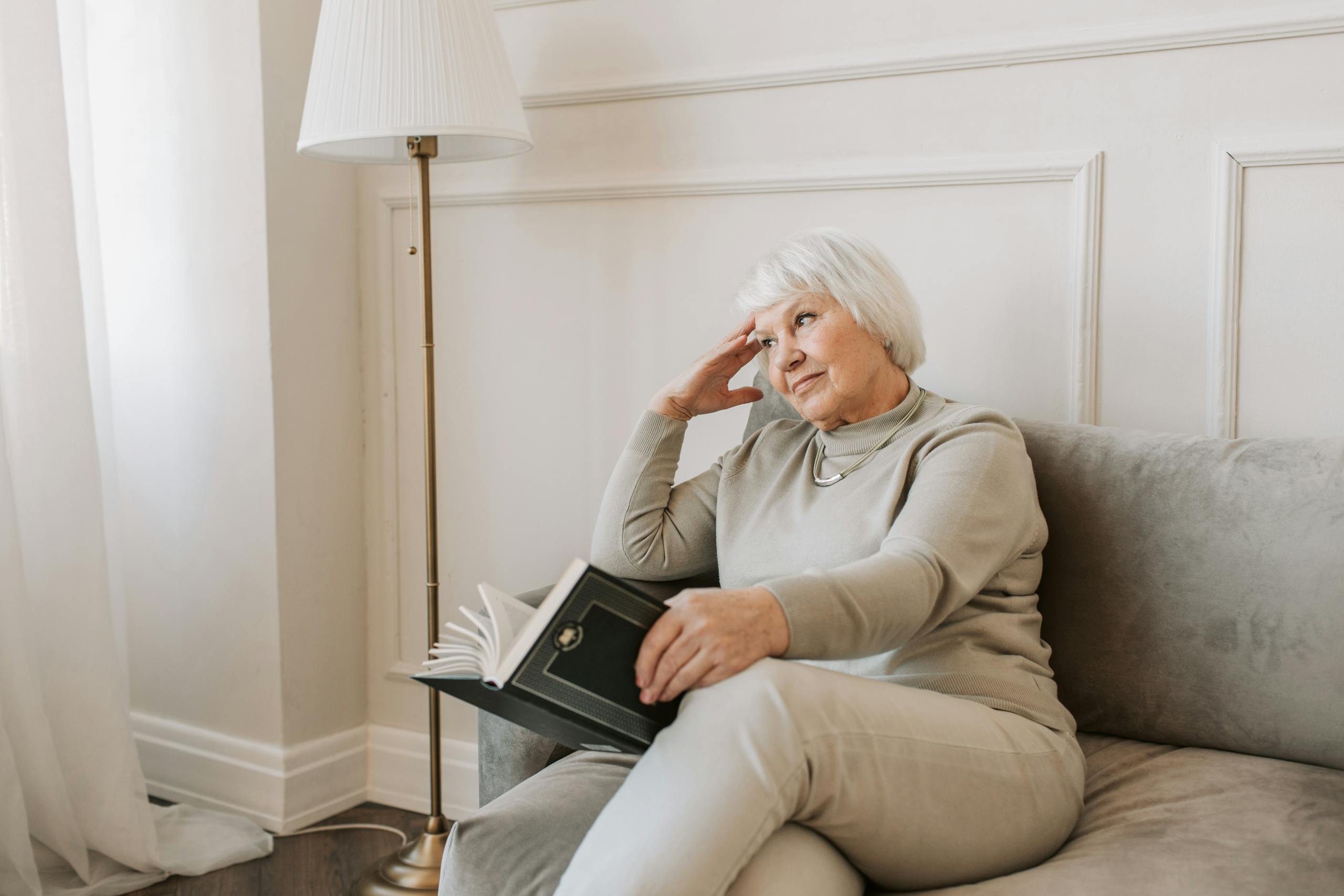 Senior woman sitting on a couch reading a book, with a thoughtful expression.