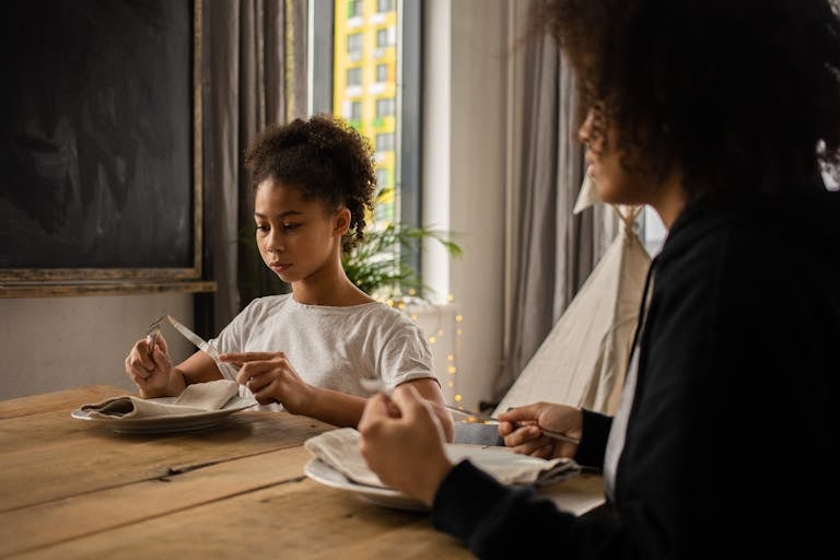 Serious African American girl with black faceless mother sitting at wooden table with plate and silverware while learning dining etiquette in room