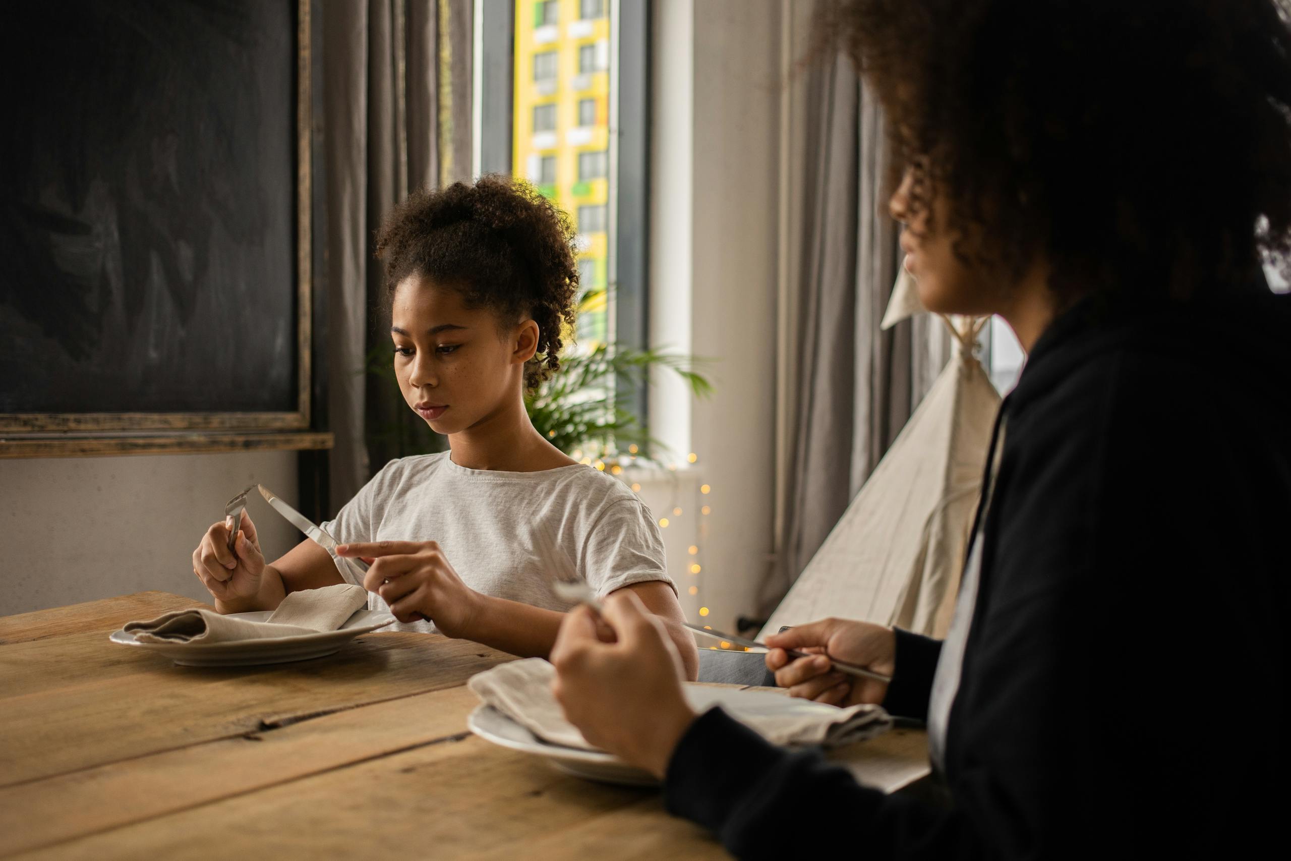 Serious African American girl with black faceless mother sitting at wooden table with plate and silverware while learning dining etiquette in room