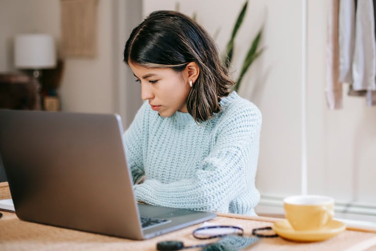 Serious young Latin American woman in casual clothes working remotely on netbook at table near mug with drink in light room with plant with green leaves and wardrobe with apparels