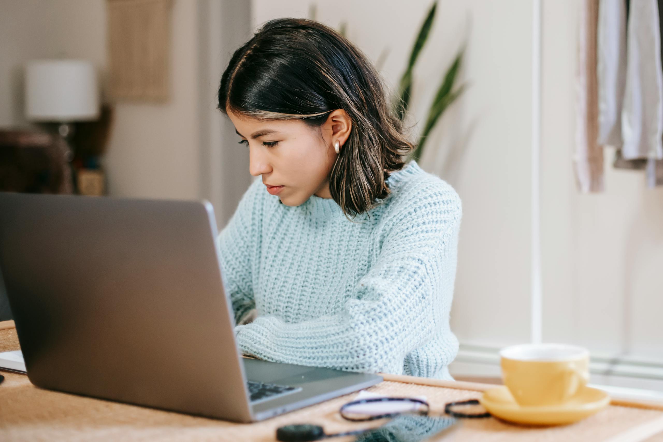 Serious young Latin American woman in casual clothes working remotely on netbook at table near mug with drink in light room with plant with green leaves and wardrobe with apparels