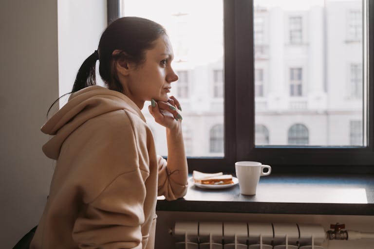 Side view of a woman in a hoodie sitting by a window, contemplating while looking outside.