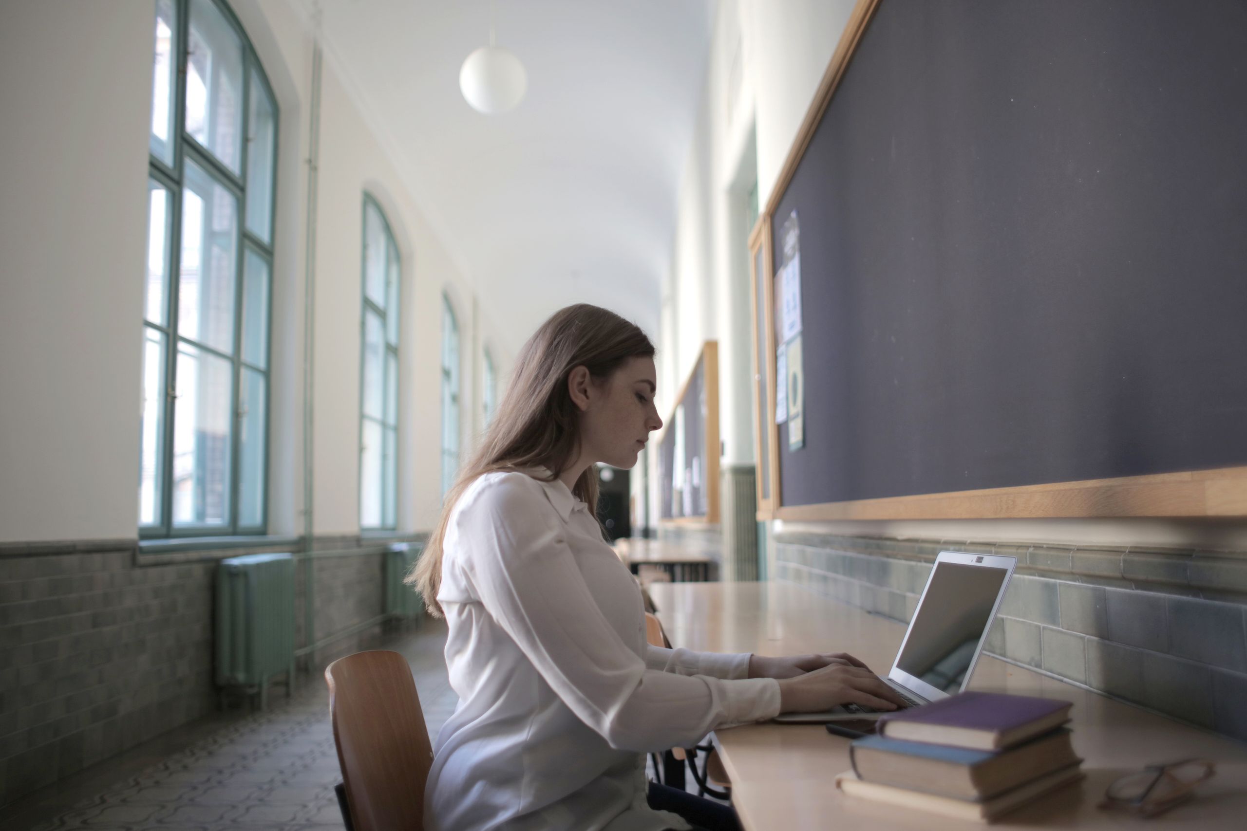 Side view of young brown haired concentrated woman typing on laptop while sitting at table with books