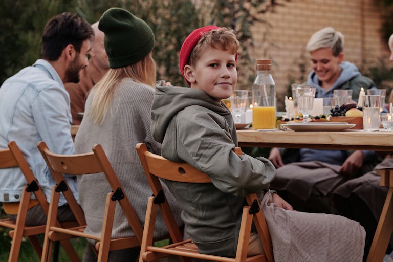 Smiling boy wearing warm clothing and with napkin across lap sitting together with relatives at wooden table during outdoor party