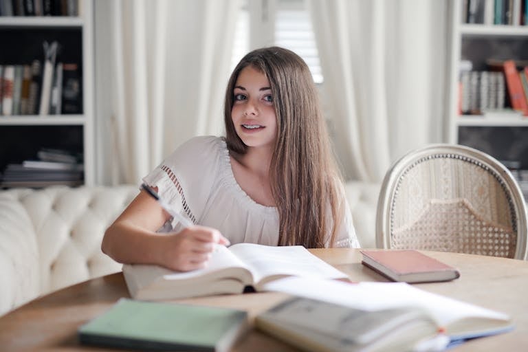 Smiling woman with long hair studying indoors at a table surrounded by books.